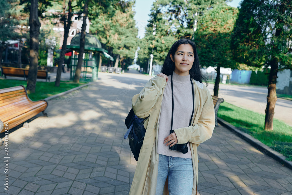 Naklejka premium Woman in light coat with camera walks through a sunny park, casual street style, greenery, and relaxed mood in an urban outdoor scene featuring bright light, breeze, and casual fashion vibe for