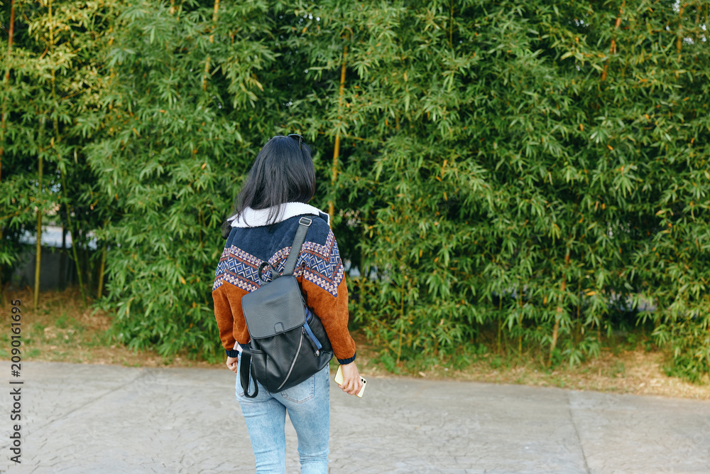 Naklejka premium Back view of a woman wearing a patterned knit sweater and backpack, strolling along a paved path beside a dense green hedge, conveying a casual outdoor commute and relaxed travel vibe
