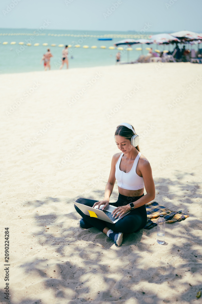 Fototapeta premium Side view of fit woman using laptop while sitting on the sand at a tropical beach