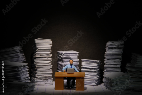 Miniature tiny people toy figure photography. Overload and work overtime concept. A businessman seat on a desk with pile of document, working until night at dark room with spotlight