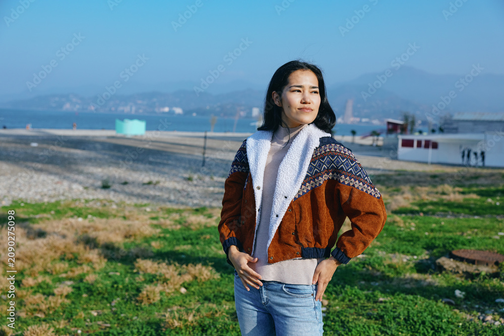 Fototapeta premium Woman standing outdoors in a burnt orange and white knit jacket with scarf, relaxed pose, exploring countryside scenery with meadow, hills and distant mountains under clear sky
