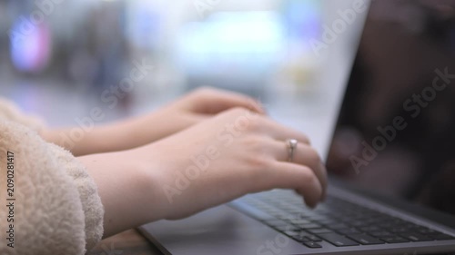 Hands type on laptop. Blurred background suggests public space. Woman wears ring. Soft lighting. No animals or plants visible