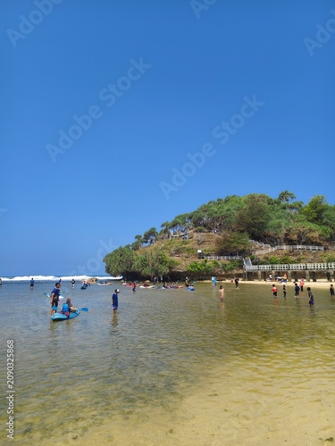 Tourists enjoying shallow ocean waters at a tropical beach with green hills under a bright blue sky