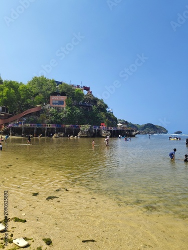 Tourists enjoying shallow ocean waters at a tropical beach with green hills under a bright blue sky