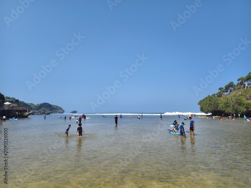 Tourists enjoying shallow ocean waters at a tropical beach with green hills under a bright blue sky