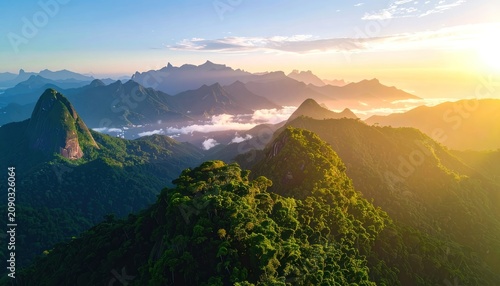 Aerial view of lush green mountains bathed in golden sunrise light, with clouds nestled in the valleys and distinct rock formations visible.