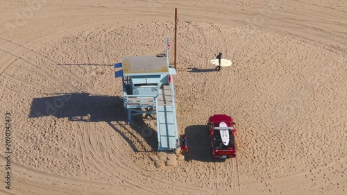 Santa Monica, California, USA - JANUARY 05, 2025: Aerial drone shot of blue lifeguard tower number 26 with red rescue truck parked nearby as surfer carries board across sandy beach.