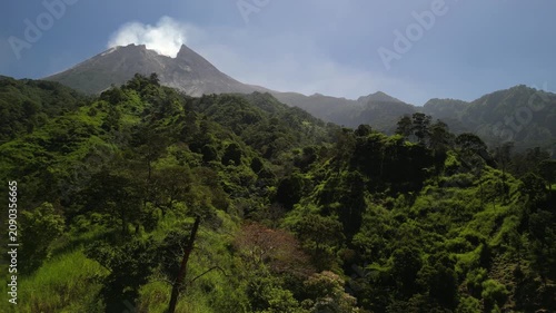 4k aerial drone video footage of Mount Merapi volcano seen from Klangon Hills emitting volcanic smoke from its summit crater, surrounded by forest mountain ridges, clear blue daytime sky.