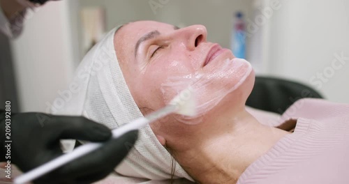 Skin mask. Facial care. Professional cosmetologist in gloves using moisturizing serum for woman client with brush in beauty salon.