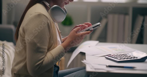 Asian woman typing on mobile phone screen while managing business budget and finance at home office.