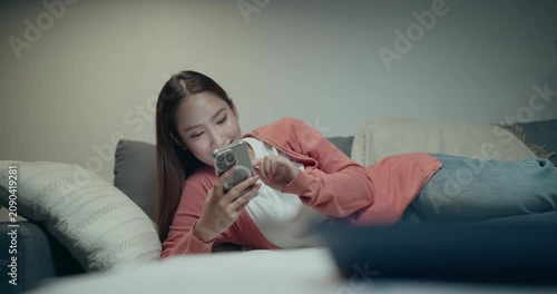 Happy Asian woman smiling and using smartphone while lying down on sofa in modern living room at night.