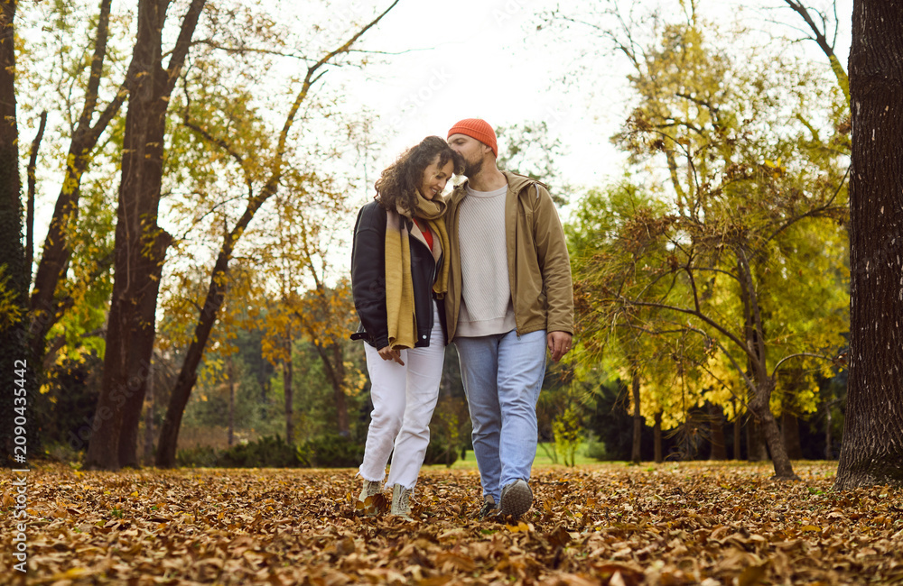Naklejka premium Couple walking in autumn park among leaves. He kisses her forehead, sharing happy warmth as they stroll together through golden foliage and nature. Love, romance, and peaceful autumn together.