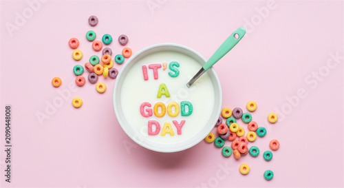 Colorful cereal rings spelling It's a Good Day in milk bowl, vibrant breakfast flat lay on pink background, positive morning message concept