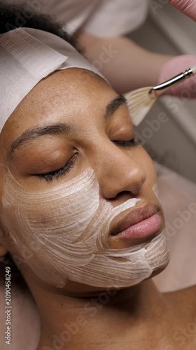 Close-up of a professional cosmetologist using a fan brush to apply a white cosmetic mask to an African American woman's face. Diverse skin care.