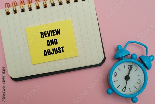 Photography Yellow sticky note with the text REVIEW AND ADJUST on a notebook next to a blue alarm clock, isolated on a pink background
