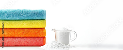 Stack of colorful folded towels next to a measuring cup with laundry detergent powder isolated on a white background