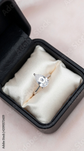A luxury jewelry box lined with ivory satin opens to reveal a custom engagement ring with a pear-cut diamond. Soft ring light creates even illumination with no harsh shadows. Background is a blurred v