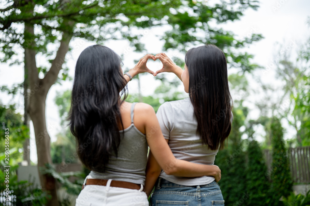 Obraz premium Rear view of two Asian women hugging each other and making a heart shape hand gesture in the park.