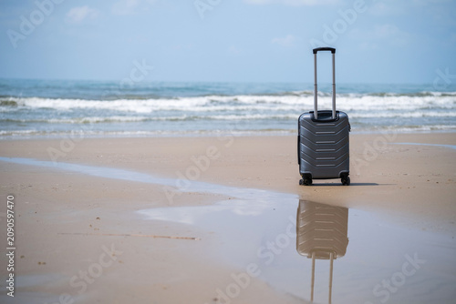A lone grey rolling suitcase standing on a sandy beach beside the calm sea with open horizon and soft cloudy sky. Vacation and travel concept. Copy space