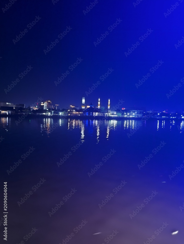 Fototapeta premium Nighttime view of a city skyline with illuminated buildings and their reflections shimmering on the calm water surface under a dark blue sky.