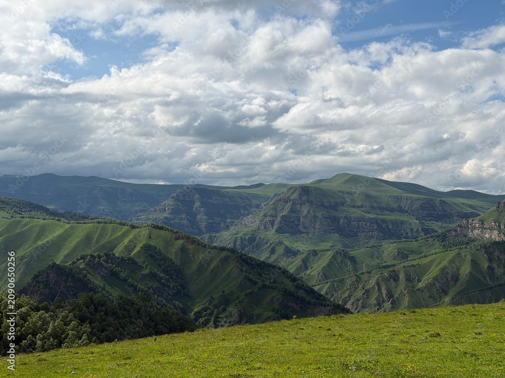 Obraz premium Lush green mountains rise under a cloudy sky, showcasing rolling hills and valleys in a breathtaking natural landscape scene