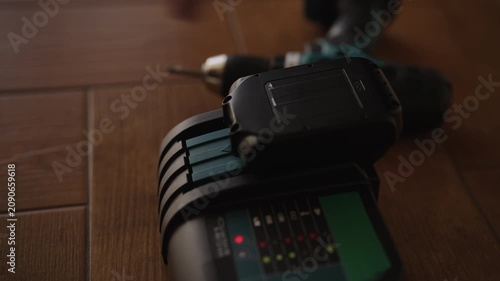 Close-up male hands inserting rechargeable battery pack into charger, with indicator lights showing charging process beginning on wooden floor. Concept of power tool maintenance.
