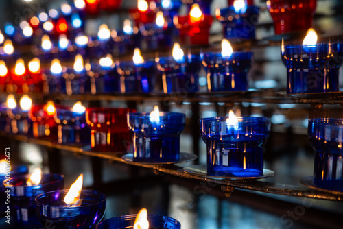 Rows of burning unlit red and blue votive candles on a stand. High angle close-up shot of white wax and wicks in glass cups, creating a repeating pattern.