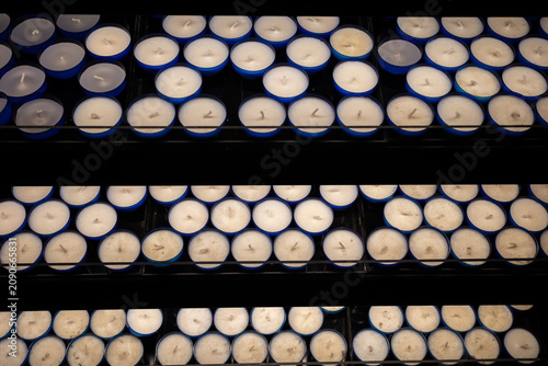 Rows of unlit red and blue votive candles on a stand. High angle close-up shot of white wax and wicks in glass cups, creating a repeating pattern.