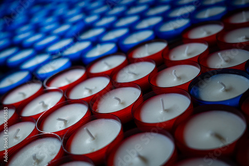 Rows of unlit red and blue votive candles on a stand. High angle close-up shot of white wax and wicks in glass cups, creating a repeating pattern.