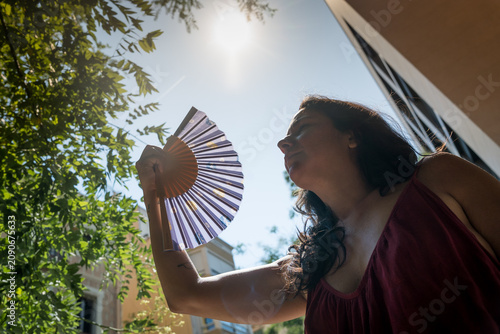 young woman outdoors during a hot summer heat wave with hand fan under the sun, cooling off fighting heat during high temperature