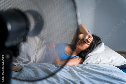 young adult woman in bed during a hot summer heat wave with fan, cooling off fighting heat at home during a heatwave and high temperature