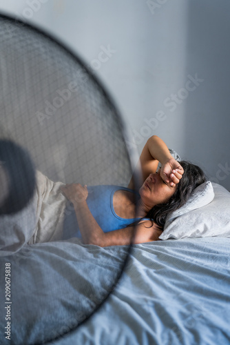 young adult woman in bed during a hot summer heat wave with fan, cooling off fighting heat at home during a heatwave and high temperature