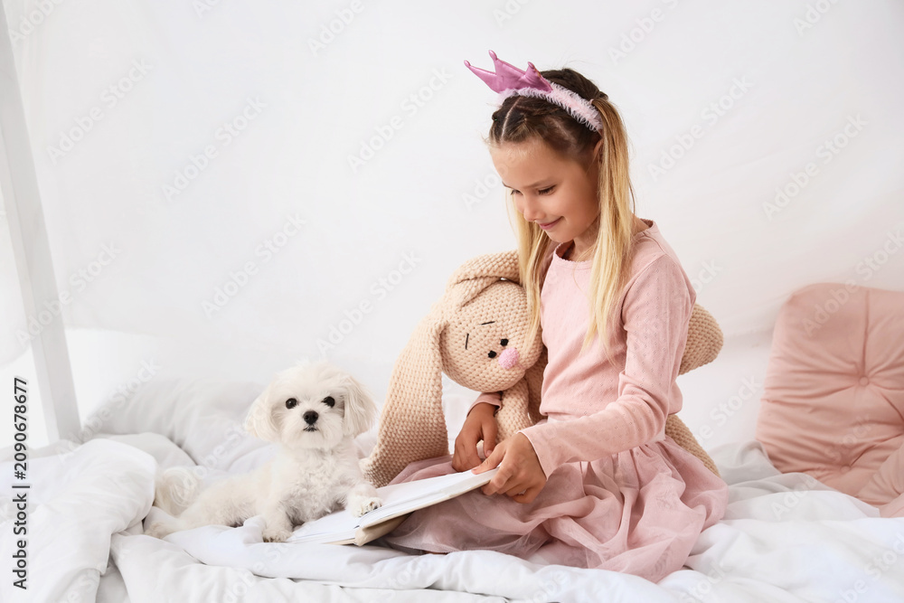 Obraz premium Cute little happy girl with Maltese dog and toy reading book on bed at home