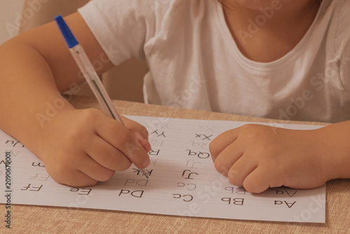 Close up horizontal shot of a child holding a pen and practicing writing english alphabet letters on a white paper sheet with dotted lines at a wooden desk