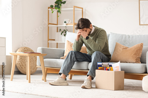 Fired young man with box of personal stuff sitting on sofa at home