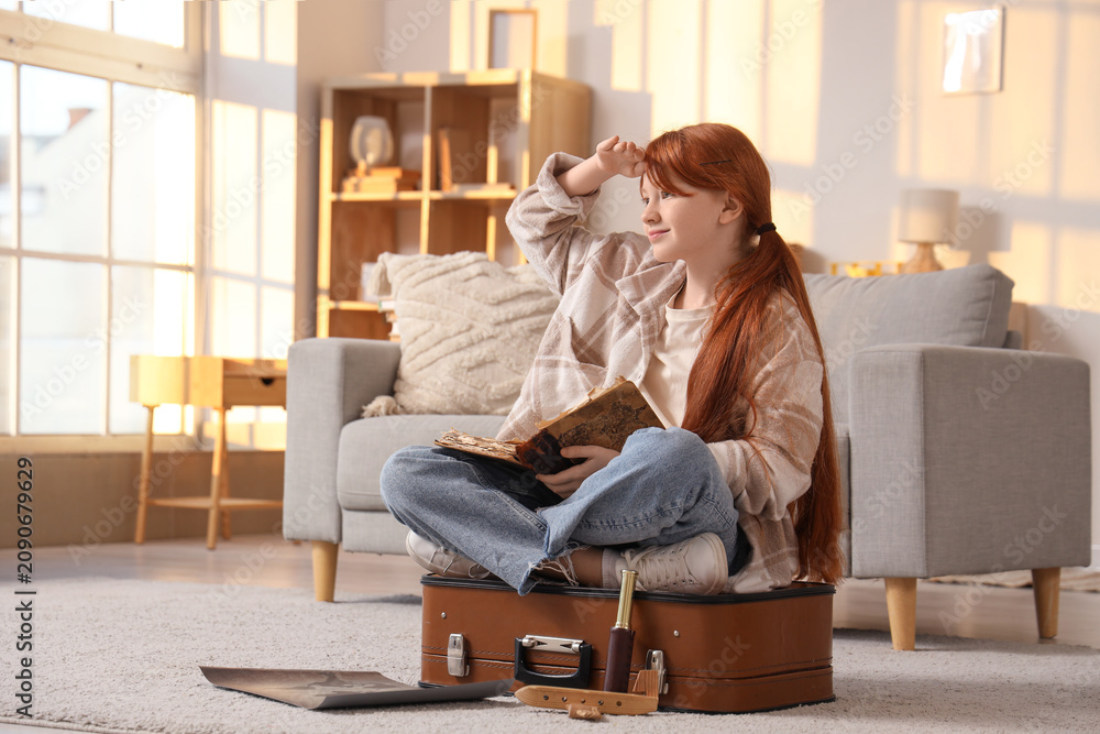 Obraz premium Cute teenage happy redhead girl with adventure book, world map and suitcase sitting at home