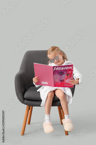 Cute little girl with bathrobe and magazine sitting in armchair on light back...