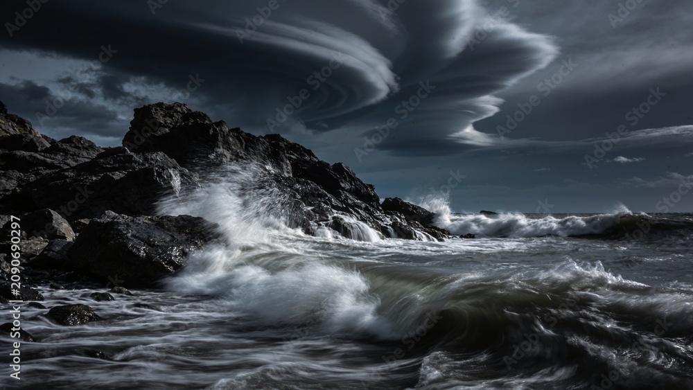 Fototapeta premium Dramatic long exposure of powerful ocean waves crashing against dark rocky cliffs beneath swirling lenticular clouds, perfect for editorial about climate resilience or coastal conservation campaigns.