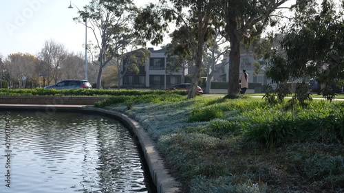 A peaceful waterfront within the master-planned residential community of Sanctuary Lakes, woman pushing a pram, residential houses and traffic in the background. Point Cook, Melbourne, Australia