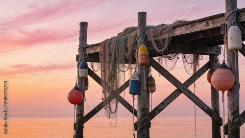 Rustic wooden pier structure adorned with hanging fishing nets and vintage buoys beneath a dramatic pink and orange sunset sky.