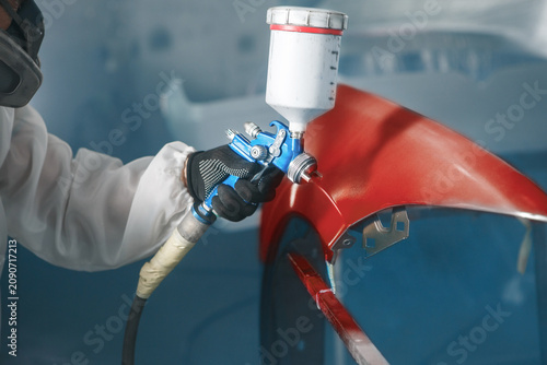 Car paint worker spraying red paint to car body element using spray gun in vehicle workshop chamber. Complex restoration of automobile body. 