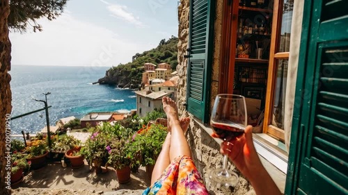 Relaxing with red wine overlooking a picturesque Italian coastal village. A person enjoys a glass of red wine on a sunny day with colorful houses and sparkling blue sea views
