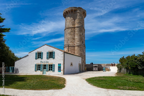 The historic old stone lighthouse tower and charming white museum building with blue shutters on a bright sunny day in Ile de Re, France.