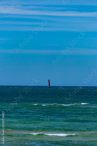 A solitary distant lighthouse stands amidst the vast blue Atlantic Ocean beneath a clear, expansive sky on a sunny day in Ile de Re, France.