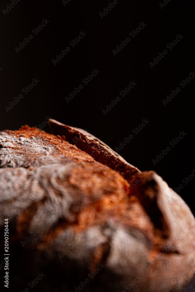 Fototapeta premium Primer plano de pan artesanal sobre fondo negro. Luz única resalta cada poro y grieta de la corteza. Estilo minimalista y dramático.