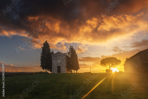 The Cappella della Madonna di Vitaleta stands silhouetted against a blazing sunset sky, with a sunstar bursting from behind a stone pine. Val d'Orcia, Tuscany, Italy.