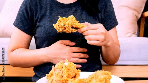 A woman holding a crispy fried chicken piece while touching her stomach on a sofa, representing stomach ache, fullness, diet awareness, and unhealthy eating habits.