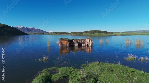 Aerial view Ancient Armenian church in Aparan Reservoir, Armenia. A solitary 5th-century stone church surrounded by calm water, with green hills and mountains in the background under a clear sky. Hist