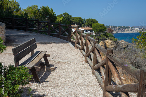 Scenic coastal pathway with wooden bench and railing overlooking the sea, surrounded by lush greenery and residential buildings in the background