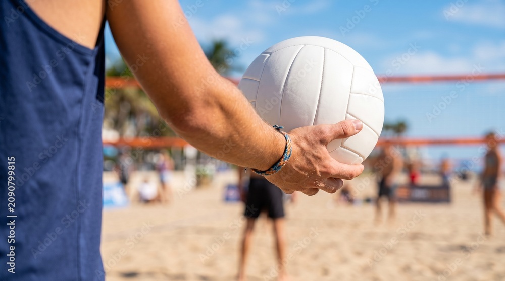 Fototapeta premium Person holding volleyball on sunny beach with players in background